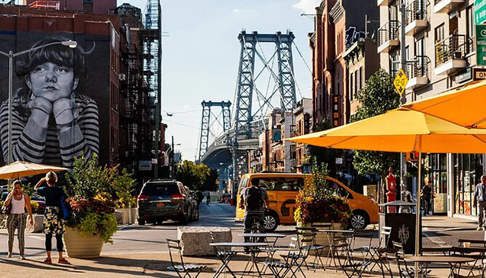 This image captures a vibrant street scene with outdoor seating under yellow umbrellas pedestrians vehicles a large mural of a girls face on a building and a distinctive bridge in the background