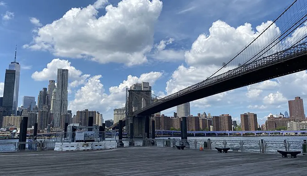 The image captures the Brooklyn Bridge extending over the East River with the Manhattan skyline in the background on a partly cloudy day