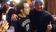 Two people are smiling for a photo, one in a Brooklyn basketball jersey and the other giving a thumbs up, with someone else taking a photo of them in the foreground.