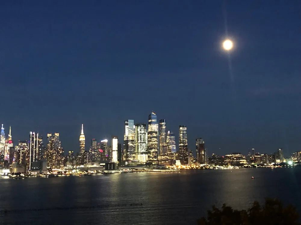 The image depicts a nighttime city skyline with bright lights reflecting on the water under a clear sky with a bright full moon overhead