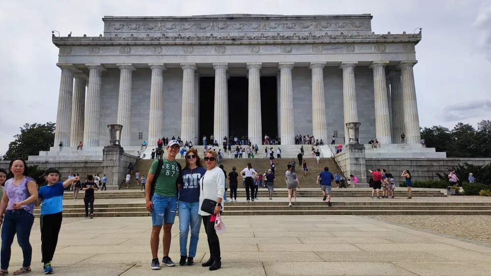 A group of people poses in front of the Lincoln Memorial a crowded historical landmark with many visitors on its steps under a partly cloudy sky