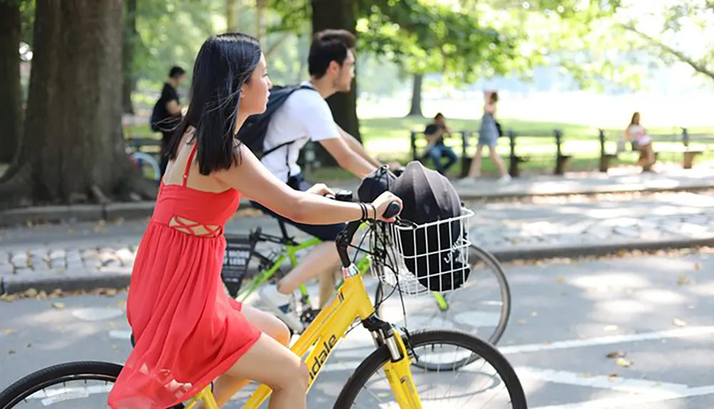 A person in a red dress is riding a yellow bicycle with a helmet in the basket along a park path while another cyclist and pedestrians are visible in the background
