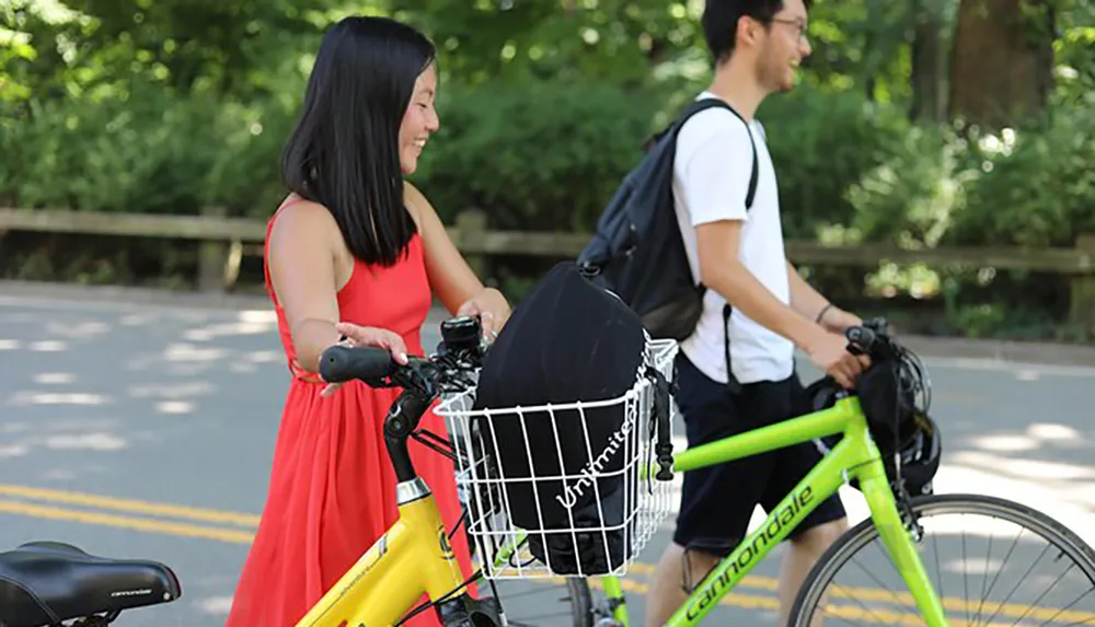 A smiling woman in a red dress pushes a yellow bicycle next to a man riding a green bicycle both on a tree-lined path