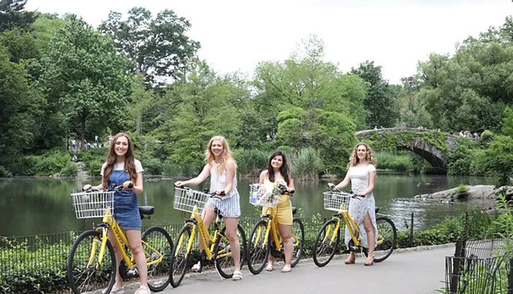 Four people are smiling and standing with bicycles in front of a picturesque pond and stone bridge surrounded by lush greenery