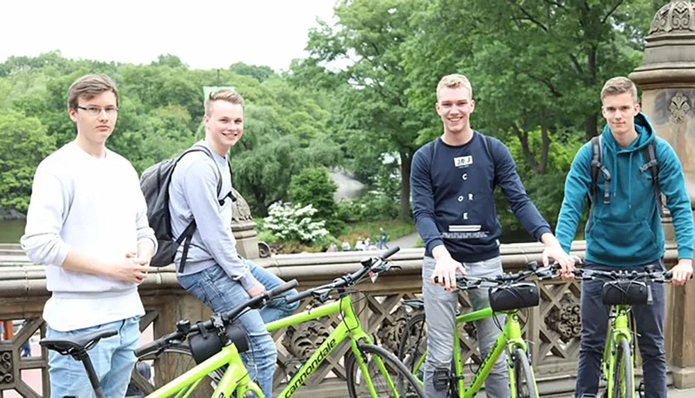 Four young men are posing with their bicycles on a bridge in a park setting