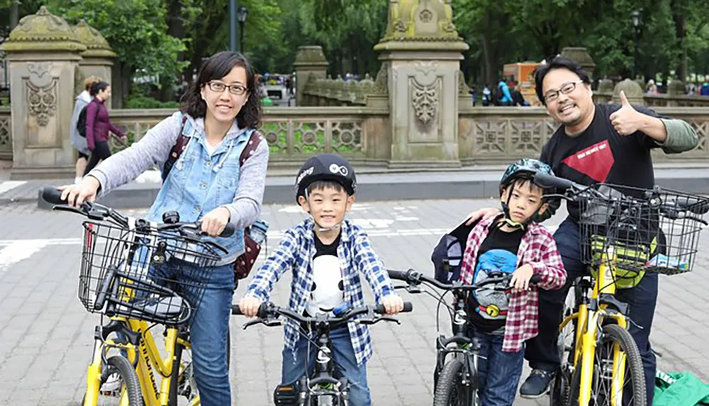 A family of four is posing for a photo with their bicycles in a park smiling and appearing to be enjoying their day out together
