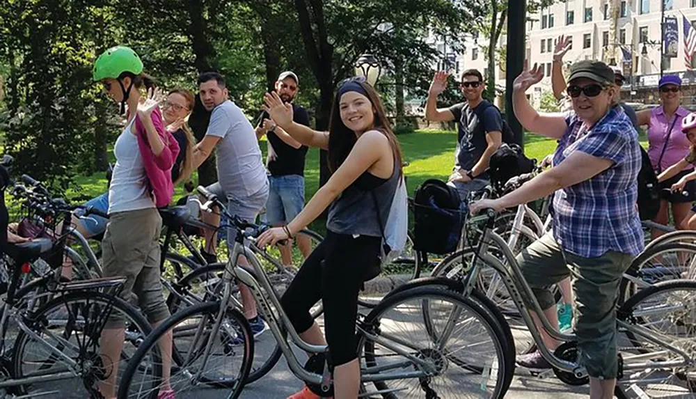 A group of cheerful people on bicycles are posing for a photo waving and smiling in a park-like setting