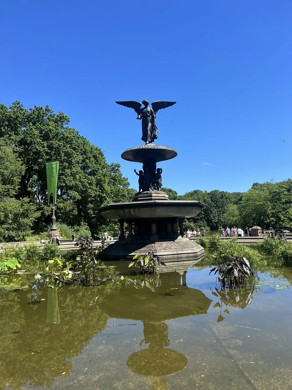 A fountain with an angel statue stands in the center of a pond surrounded by greenery under a clear blue sky