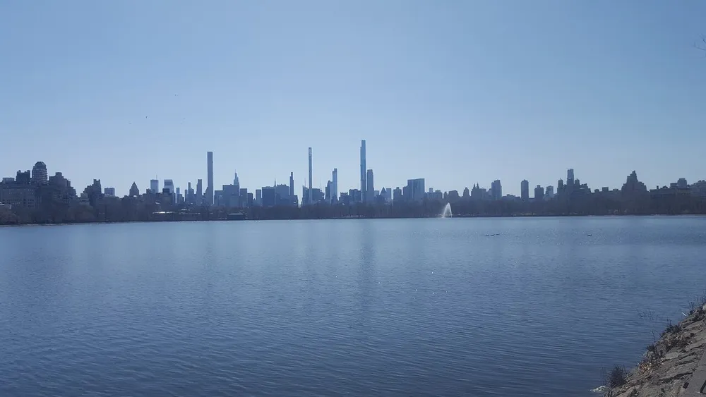 The image features a tranquil reservoir with a fountain in the foreground and a backdrop of a dense urban skyline under a clear blue sky
