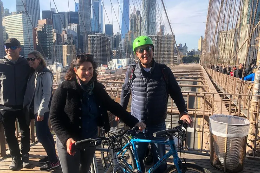 A smiling couple is posing with a bicycle on a crowded pedestrian walkway of the Brooklyn Bridge with the Manhattan skyline in the background