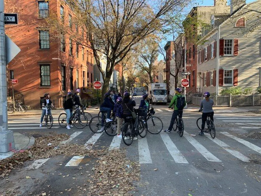 A group of cyclists is waiting at a pedestrian crossing on a tree-lined street with brick buildings on a sunny day