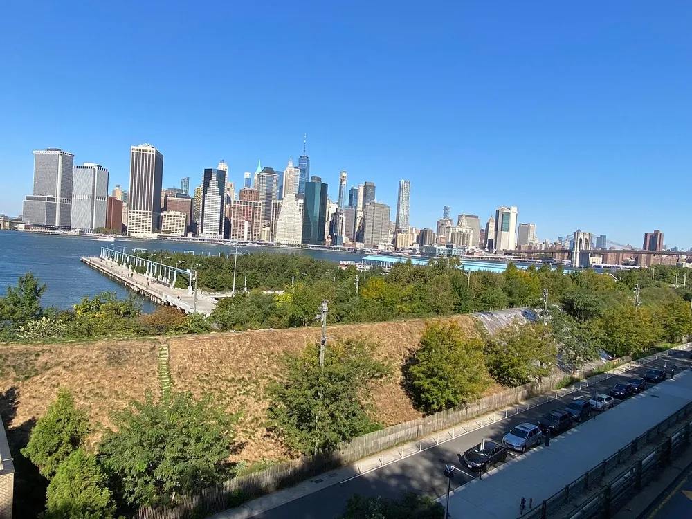 The image shows a panoramic view of a city skyline across a river with a mix of high-rise buildings under a clear blue sky