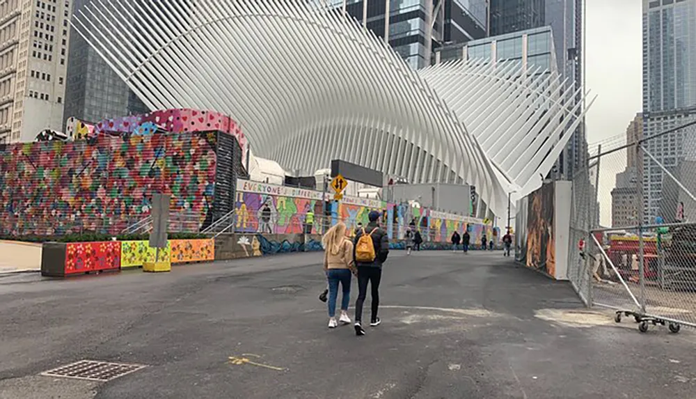 Two people walk towards the distinctive white winged structure of the Oculus in New York City with colorful street art visible on the barriers to the left