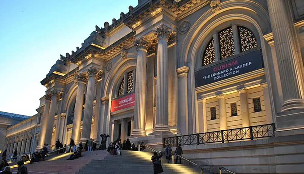 The image shows the exterior of an elegant building featuring classical architecture with banners indicating an art exhibition and multiple people sitting and walking on its steps during what appears to be early evening