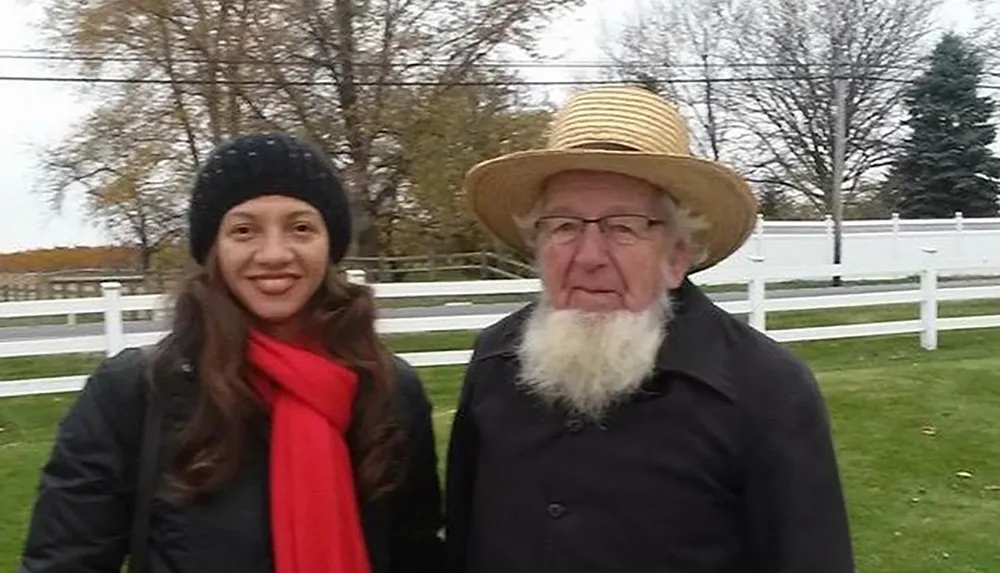 The image shows an elderly man with a straw hat and a long white beard standing next to a smiling younger woman wearing a black beanie and a red scarf with a white fence and trees in the background