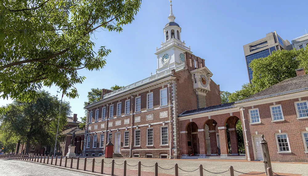 The image shows the historic Independence Hall in Philadelphia on a clear day with its distinctive brick faade and clock tower surrounded by trees and a cobblestone street in the foreground