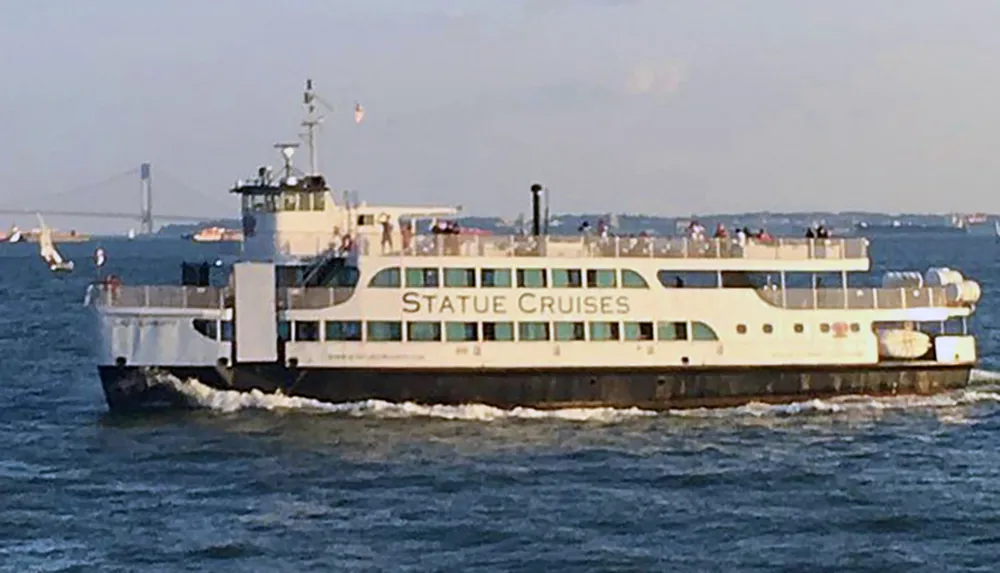 The image shows a Statue Cruises ferry boat moving on water with passengers visible on its decks