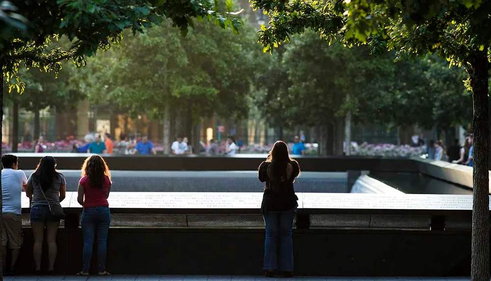 Visitors stand contemplatively before the sunlit tranquil reflective pool of a memorial surrounded by trees