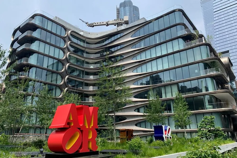 The image shows a modern wavy architectural building in the background with the bold red and yellow sculpture spelling AMOR in the foreground all amidst green foliage