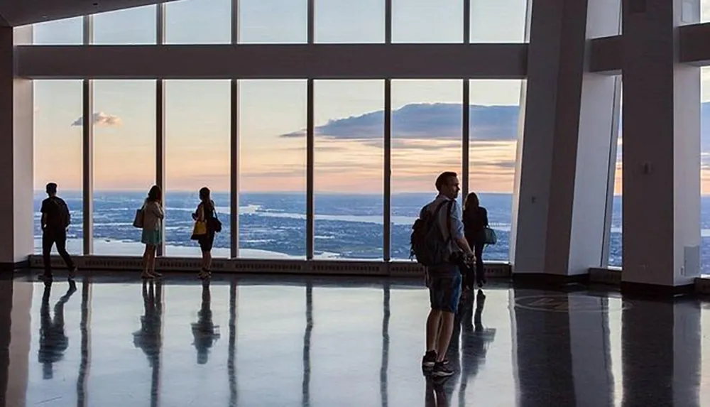 Visitors stand silhouetted against large windows offering a view of the sky and horizon at dusk in a high-rise buildings observation deck