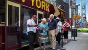 A tour guide is assisting a couple with a map in front of a sightseeing bus in a bustling city area, likely offering them information about local attractions.