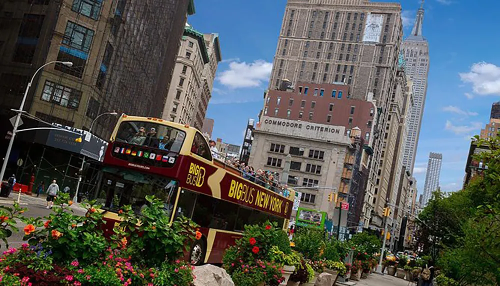 A red double-decker tour bus filled with passengers travels through a bustling urban street lined with tall buildings and vibrant flowers with the Empire State Building visible in the background
