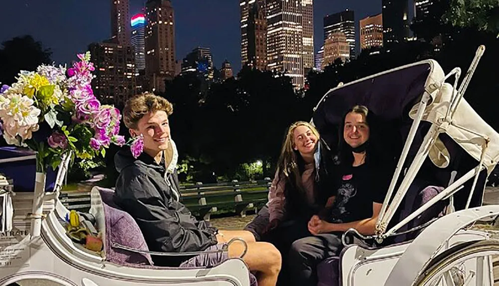 Three individuals are smiling and enjoying a horse-drawn carriage ride at night with a backdrop of illuminated city buildings