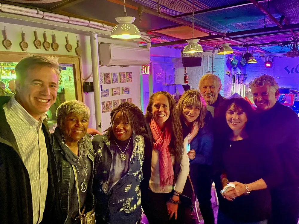 A group of eight smiling individuals pose for a photo in a room with vibrant purple lighting and musical equipment in the background