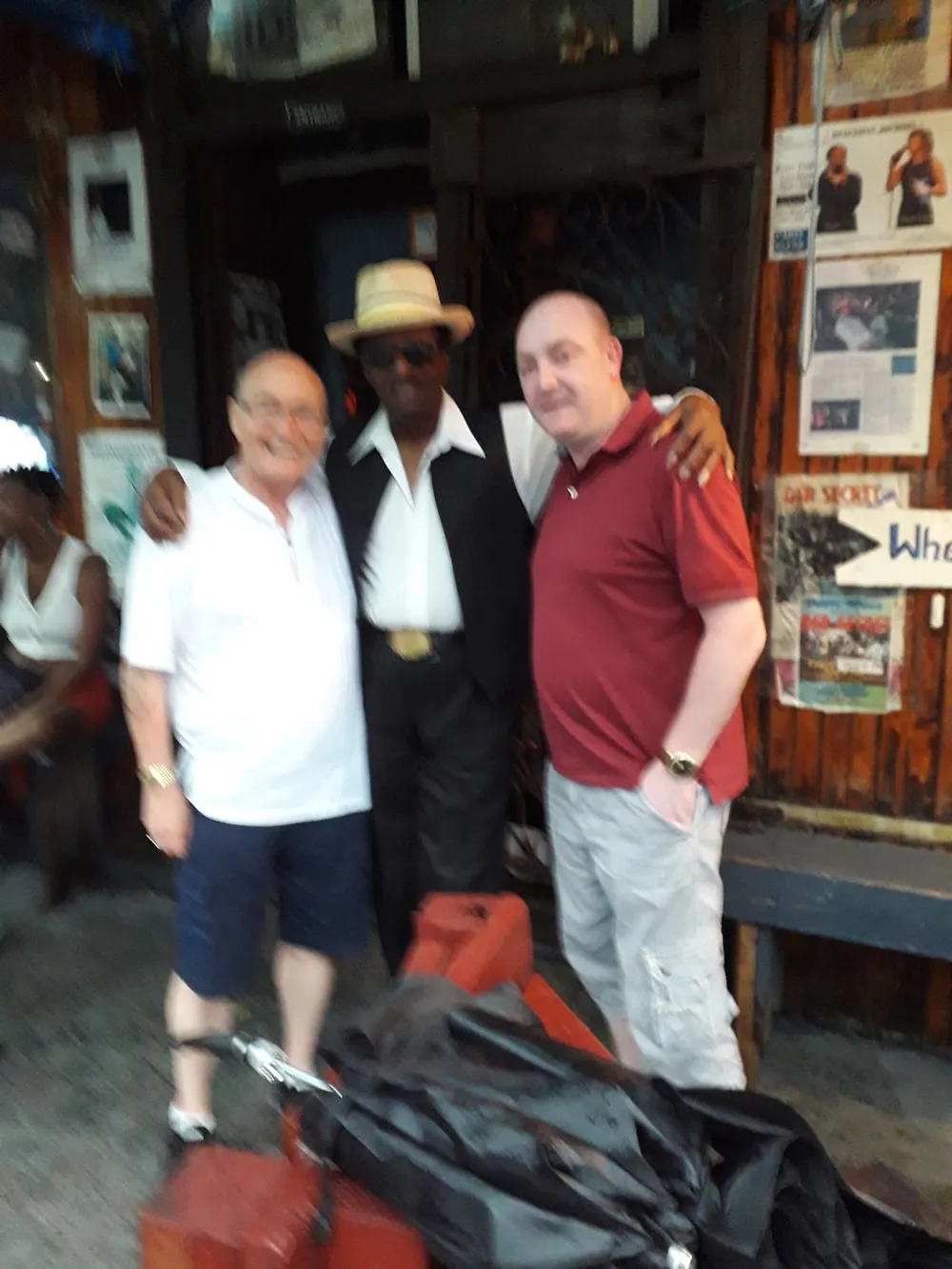 The image is a blurry photo of three men posing together two on the sides wearing casual clothes and another in the center with a hat and sunglasses in an environment that appears to be a bar or music club evidenced by posters and dark interior
