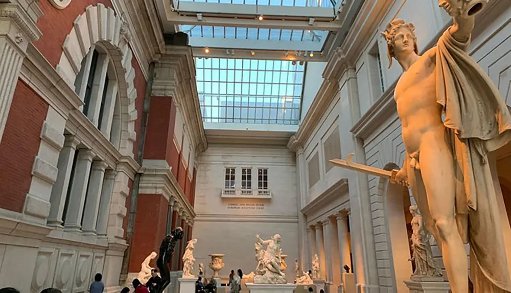 The image shows a spacious gallery with classical sculptures on display visitors admiring the art and a large window allowing natural light to fill the room