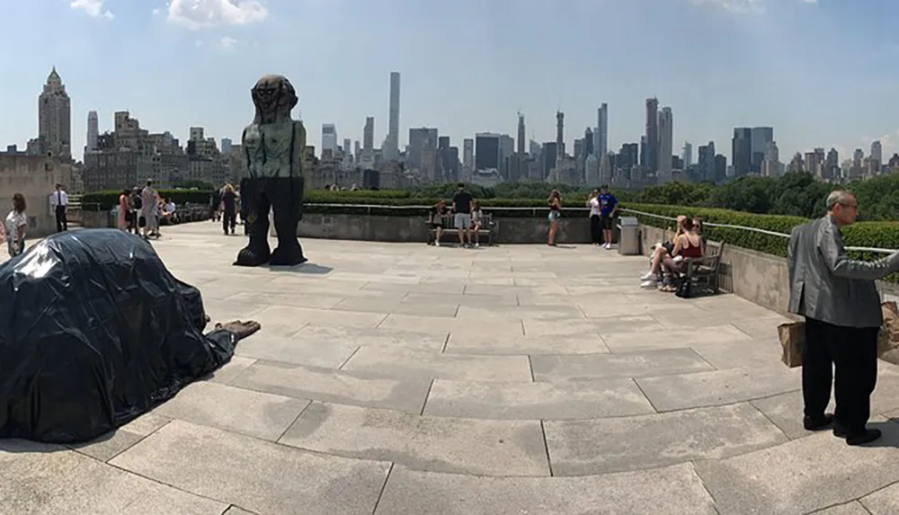 The image shows visitors at an outdoor exhibition space overlooking a cityscape with sculptures on display and a city park in the background