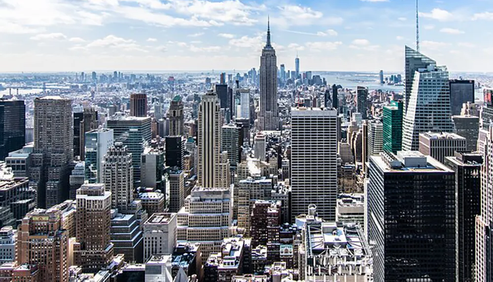 An aerial view of the Manhattan skyline with the Empire State Building in the center set against a backdrop of blue skies and scattered clouds