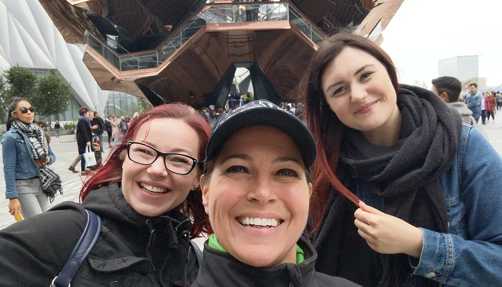 Three smiling women are taking a selfie in front of a uniquely designed modern building with a crowd of people in the background