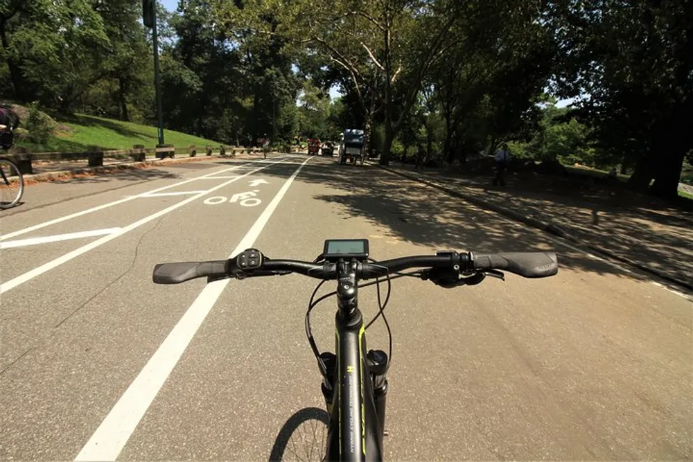 A person is cycling on a paved road through a park with the cyclists perspective shown from behind the handlebars