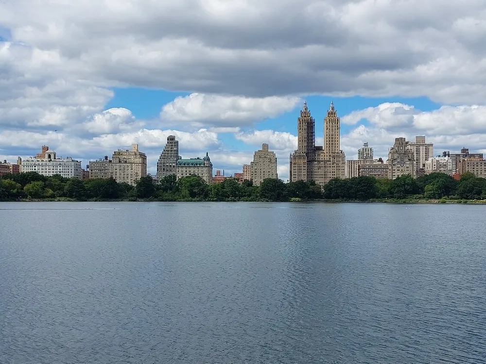 The image shows a serene body of water in the foreground with a backdrop of an urban skyline dotted by clouds in the sky