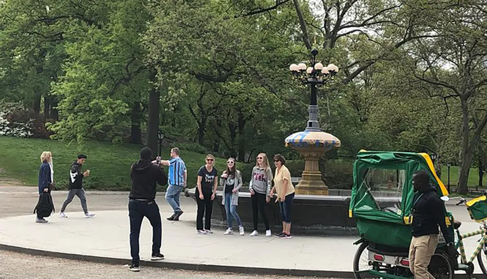 A group of people are posing for a photo taken by a man in a black hoodie in what appears to be a park with a lamppost in the background and a pedicab on the right