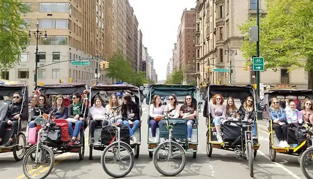 A line of pedicabs each carrying passengers is trailing down an urban street with trees and apartment buildings on either side