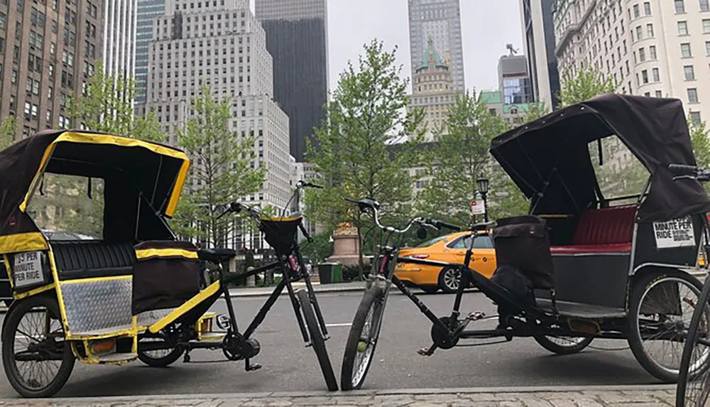 Two pedicabs are parked on a city street with trees and a yellow taxi in the background