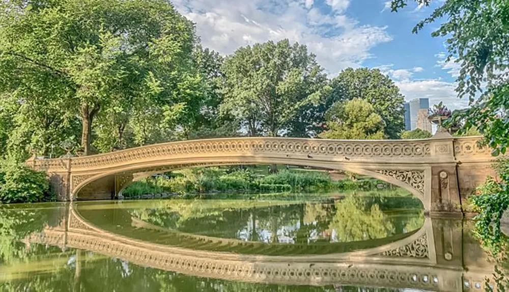 The image showcases an ornate bridge with an elaborate balustrade reflected in the calm waters of a pond surrounded by lush greenery with the hint of urban buildings in the background
