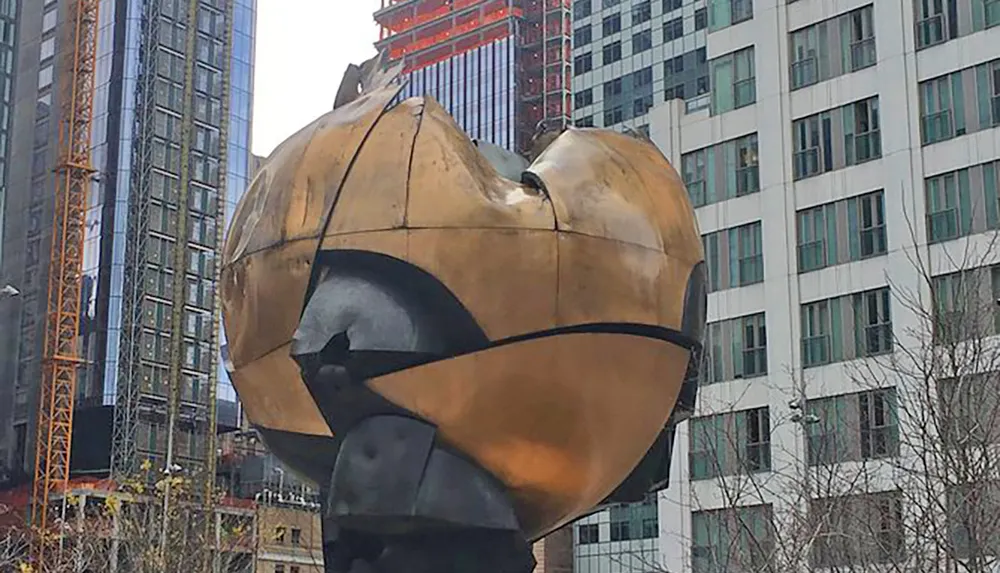 The image shows a large bronze sphere-shaped sculpture with a gash through it situated in an urban setting with high-rise buildings in the background