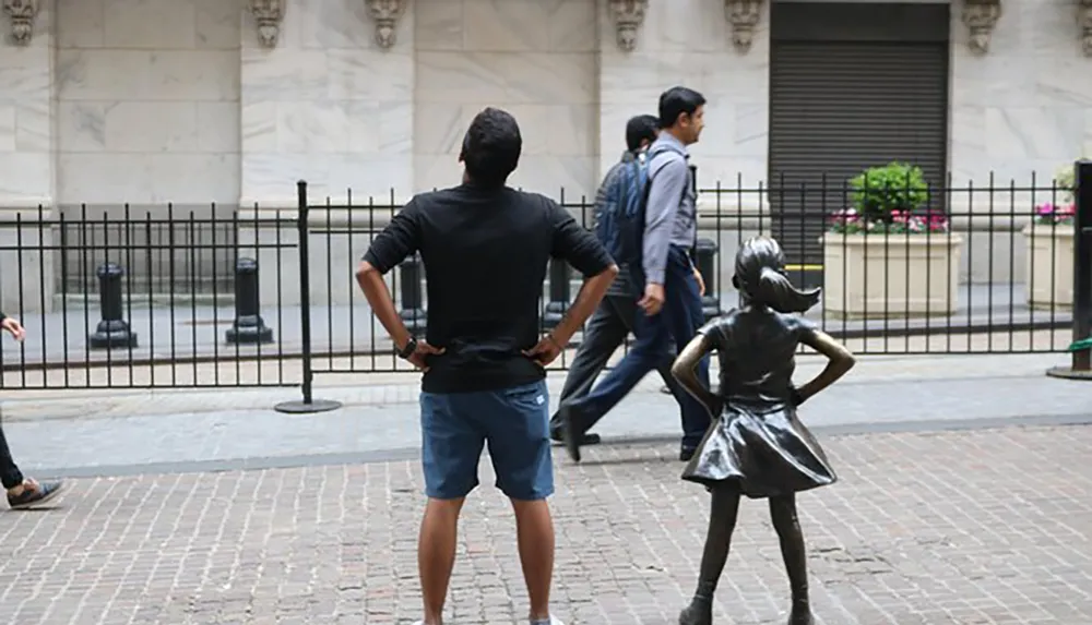 A man mimics the stance of the Fearless Girl statue standing with hands on his hips and legs apart facing away from the camera