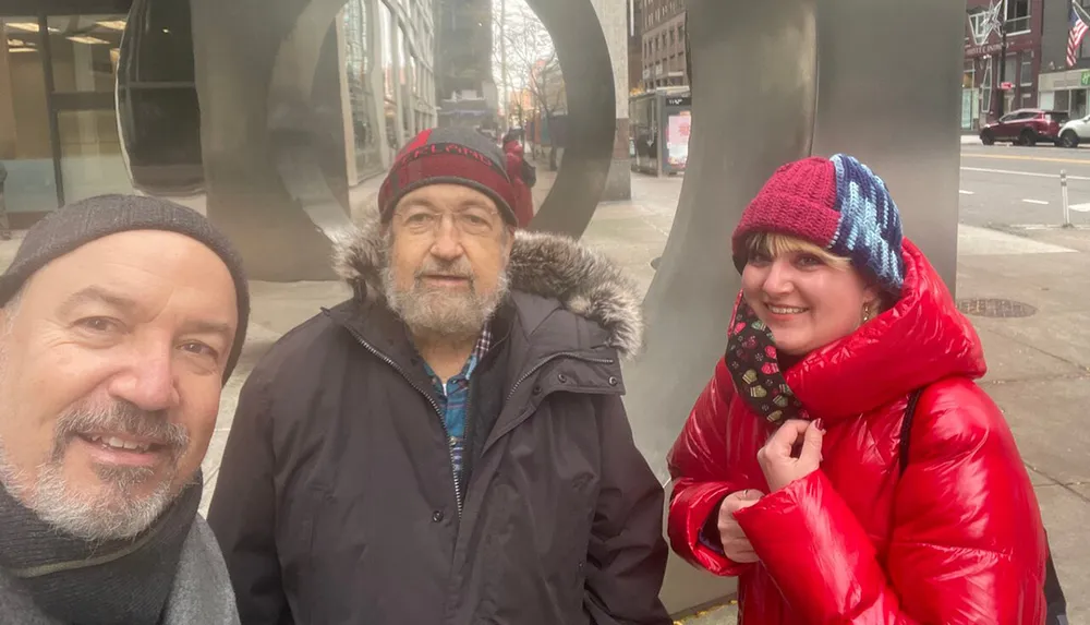 Three people are smiling for a selfie on a city street dressed in warm clothes with a large abstract metal sculpture in the background