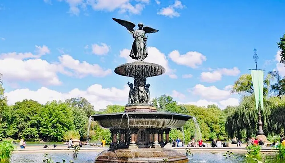 The image shows a grand fountain with water cascading from multiple tiers and a statue at the top set in a sunny park with people leisurely gathered around