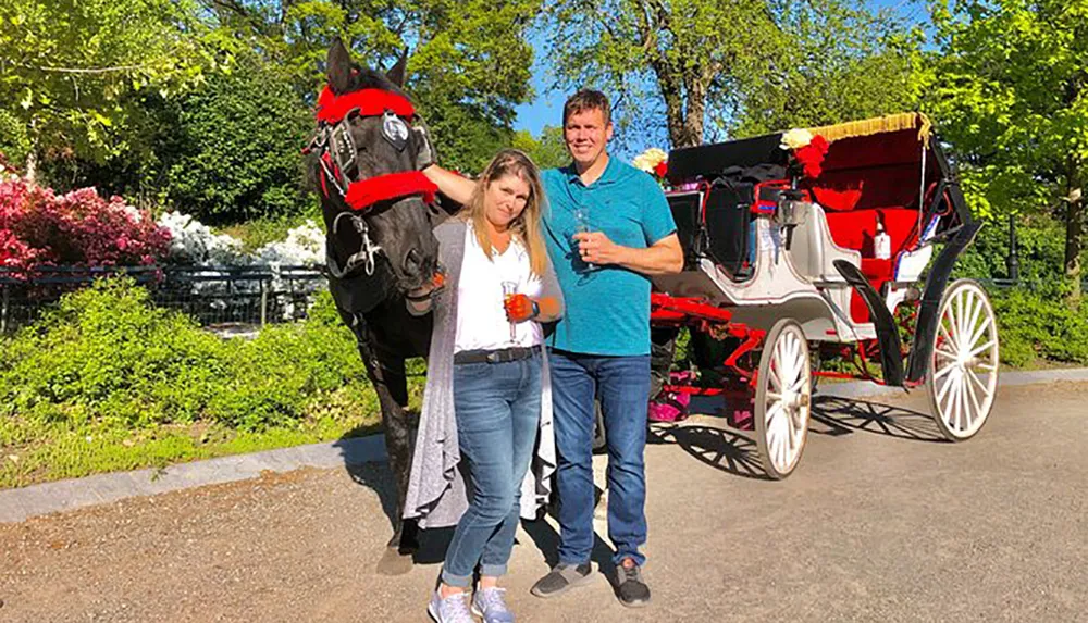 A couple smiles for the camera while each holding a beverage and standing next to a horse that is harnessed to an ornate red and white carriage with lush vegetation in the background
