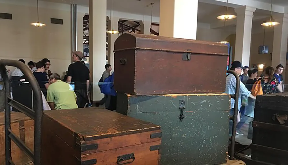 The image shows a collection of vintage trunks displayed in a room with people milling about in the background suggesting a museum or exhibition setting