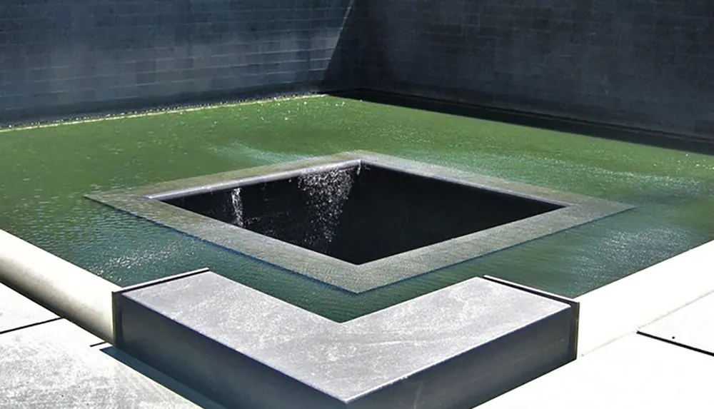 The image shows an architectural water feature with a reflective pool and a cascading waterfall effect into a lower square basin creating a serene and contemplative setting