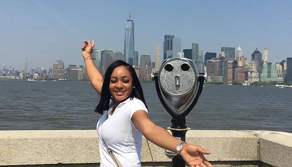A person is posing with their arm extended simulating touching the top of the One World Trade Center across the river with the New York City skyline in the background