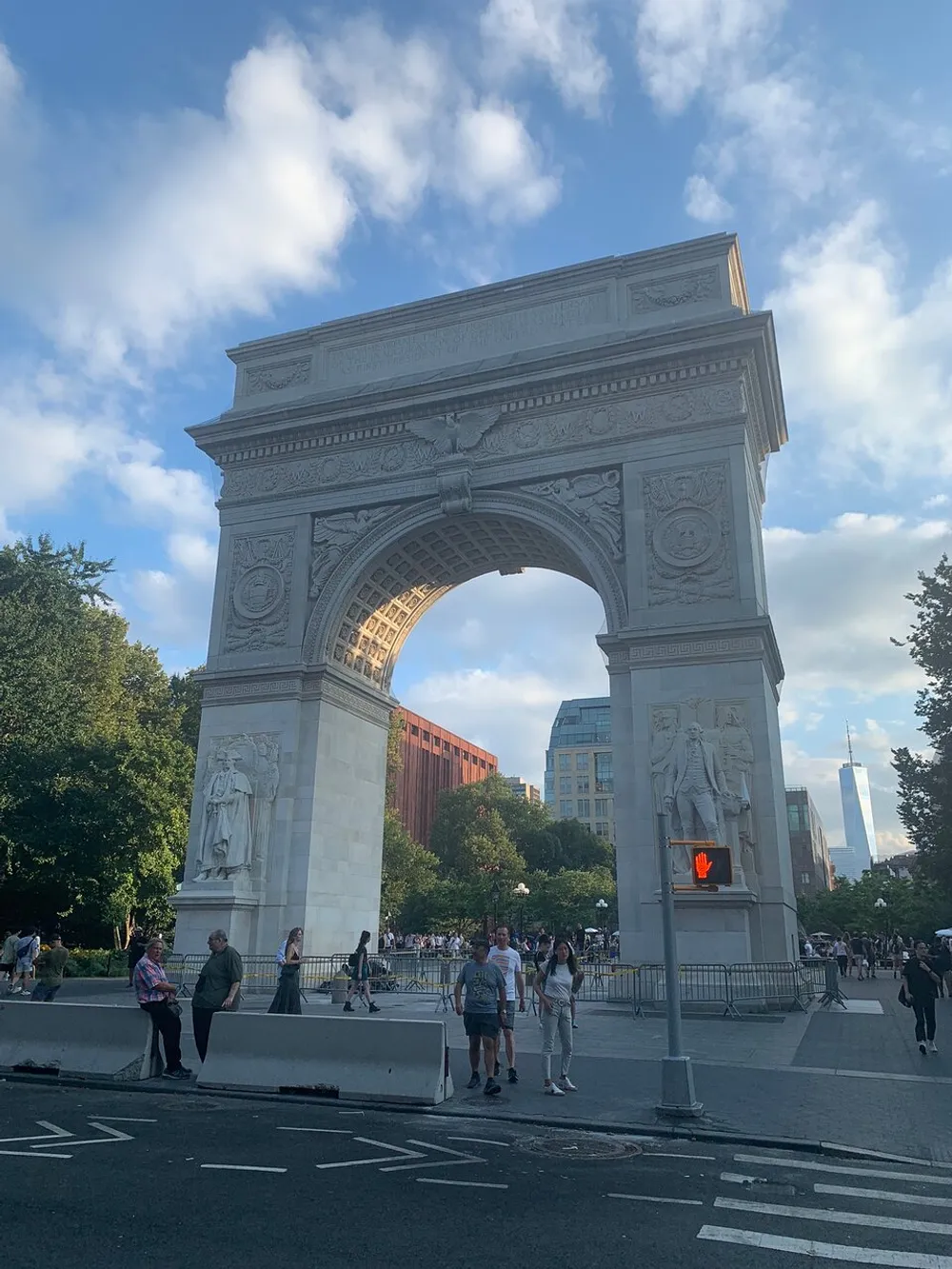 The image shows the Washington Square Arch in New York with people walking around and the One World Trade Center visible in the background under a partly cloudy sky
