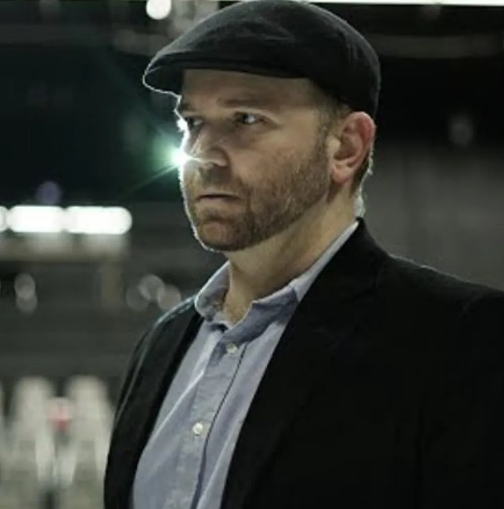A man in a flat cap and jacket casts an intense gaze over his shoulder with a dimly lit background punctuated by a bright light source