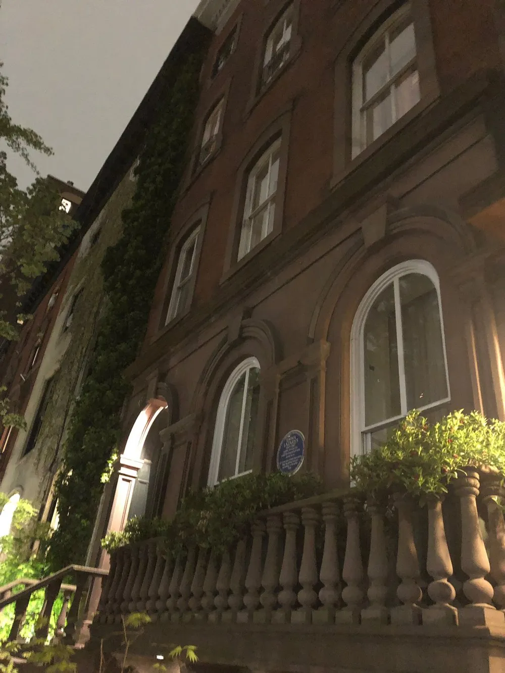 This image shows a nighttime view of a brownstone building with large arched windows a blue commemorative plaque and plants decorating the entrance staircase